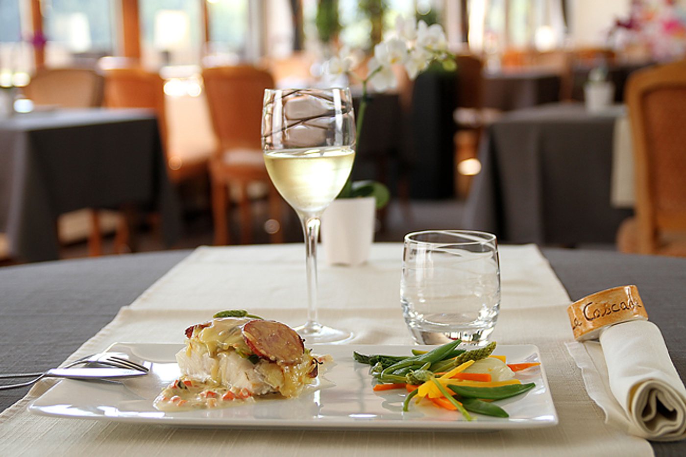 Sur une table de restaurant dressée, une assiette carrée de poisson nappé de sauce et de légumes verts accompagne un verre de vin blanc et un verre d'eau.