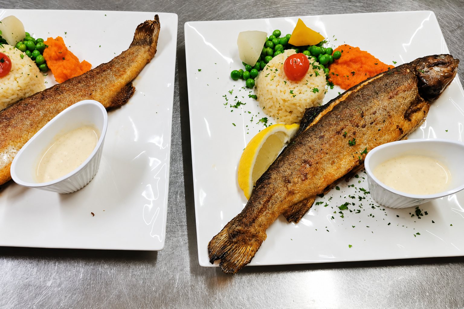 Deux assiettes carrées blanches présentent une truite entière grillée accompagnée de riz, petits pois, purée de carotte, citron et ramequin de sauce crémeuse, posées sur un plan de travail en inox.