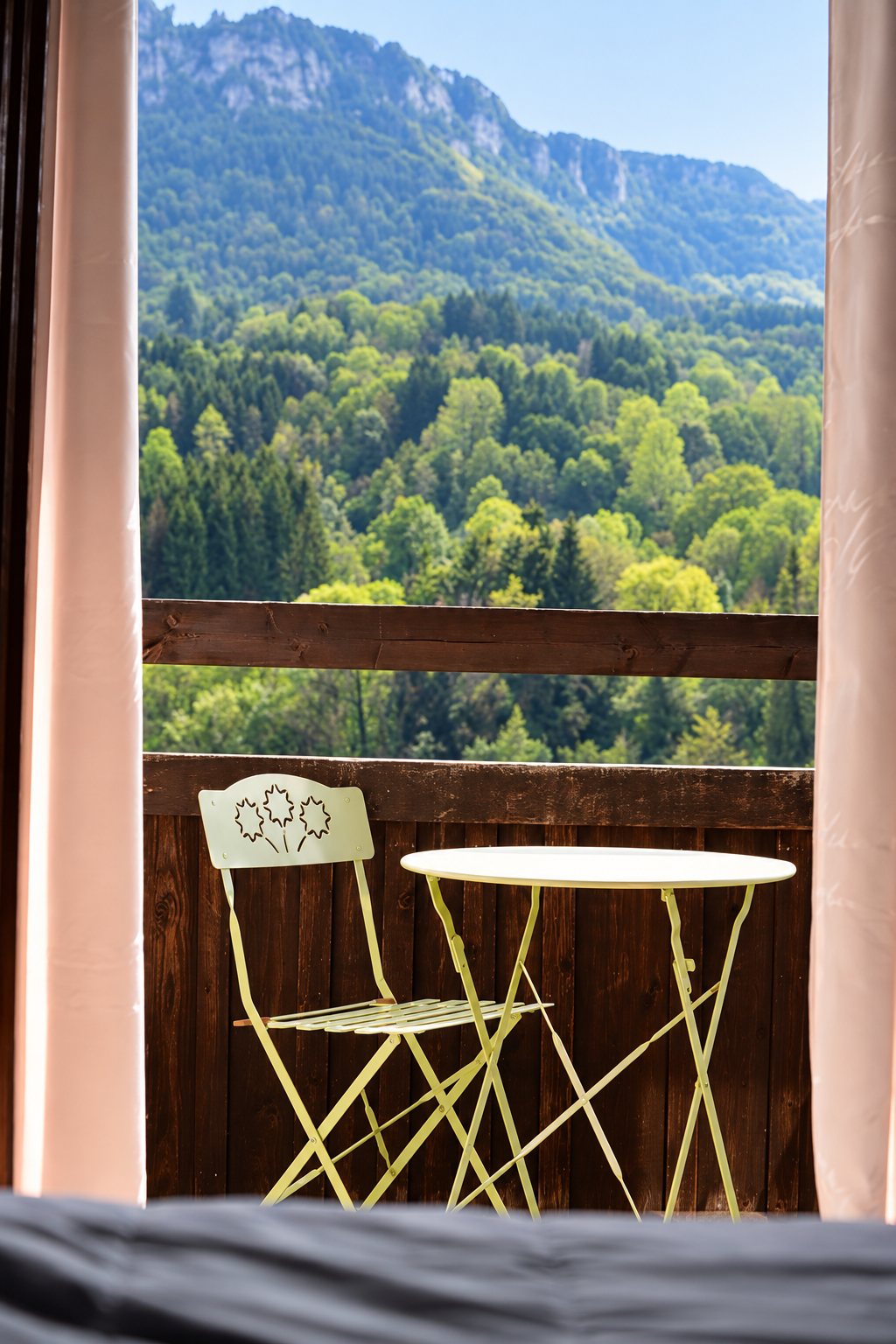 Depuis une chambre, vue entre deux rideaux sur un balcon en bois avec une table et une chaise pliantes vert pâle, face à une montagne boisée.