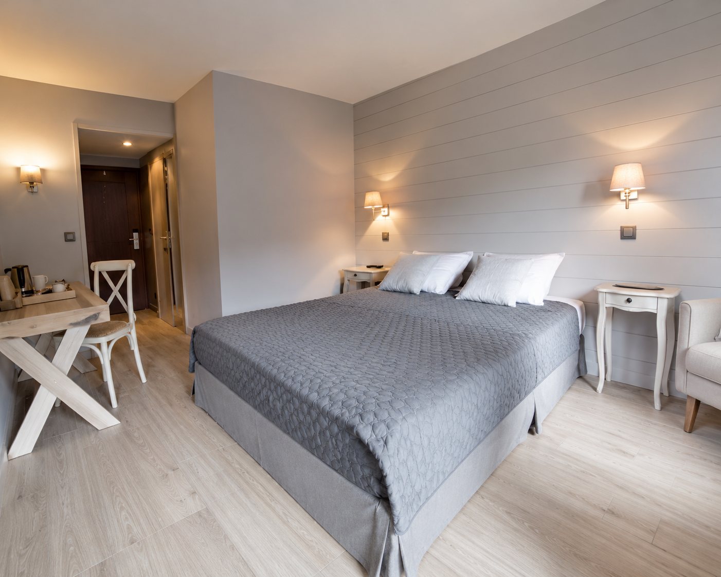 Chambre lumineuse aux tons neutres avec un grand lit double, une chaise et une table de chevet près d'une fenêtre.