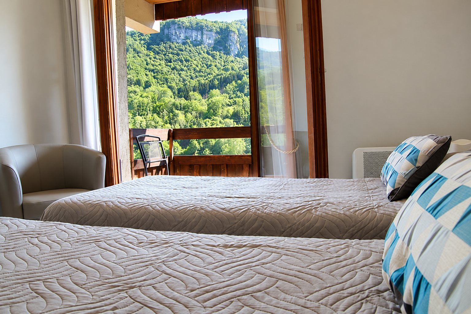 Chambre avec deux lits aux couvre-lits matelassés et fauteuil gris, dont la porte-fenêtre ouvre sur un balcon avec vue sur une falaise et une forêt verdoyante.
