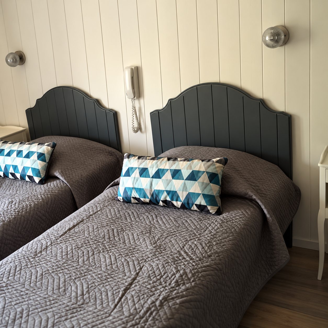 Chambre avec deux lits simples aux têtes de lit grises, couvre-lits gris à motifs et coussins décoratifs bleus et blancs, devant un mur lambrissé clair.