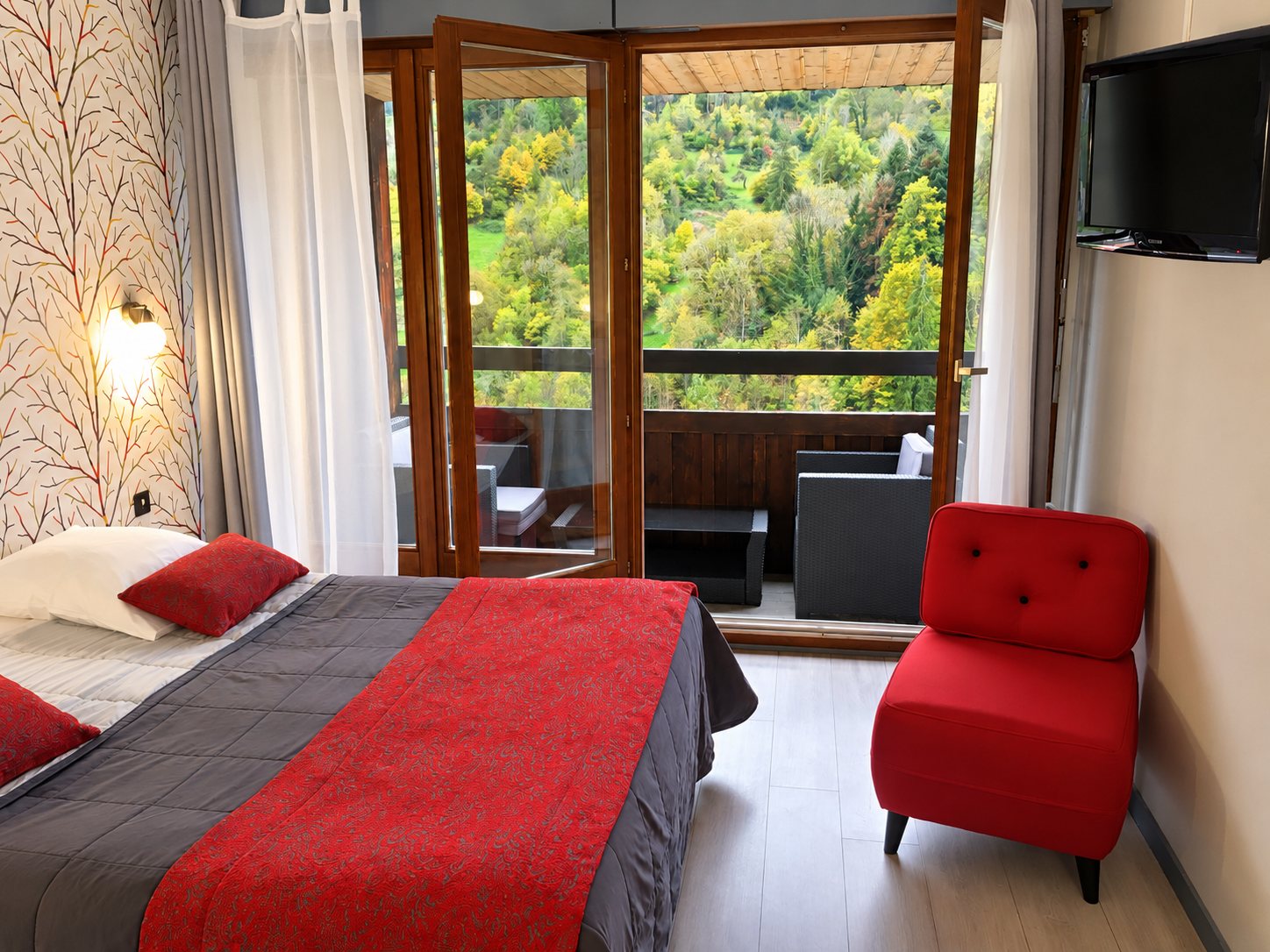 Chambre d'hôtel avec un grand lit à plaid rouge et gris, un fauteuil rouge et une porte-fenêtre ouvrant sur un balcon face à une colline boisée.