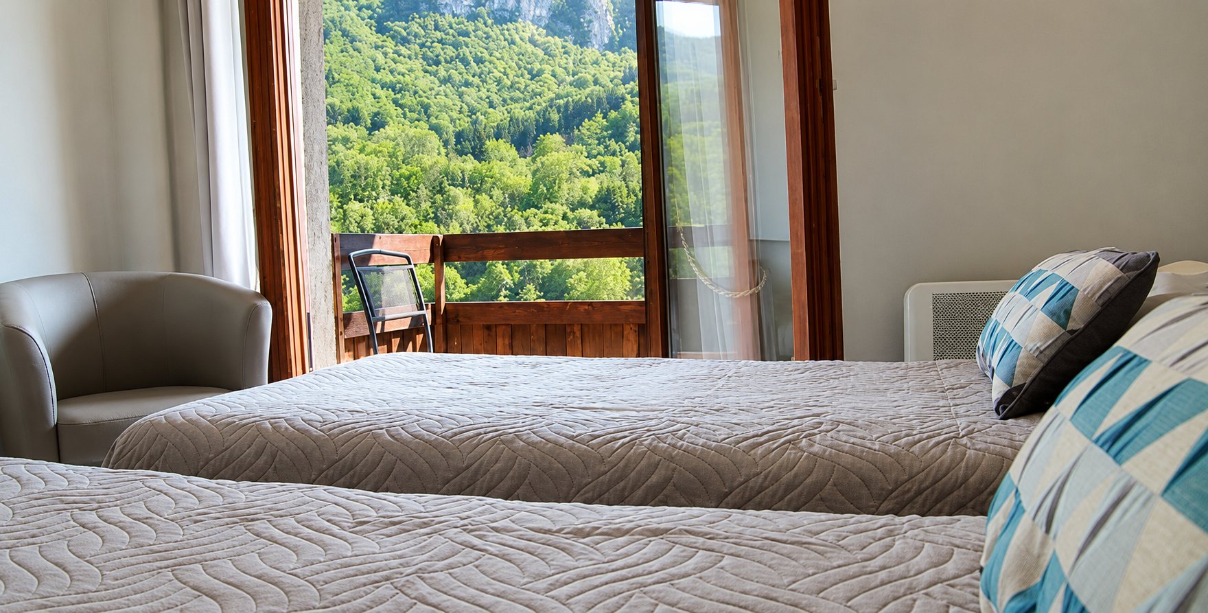 Chambre d'hôtel lumineuse avec deux lits couverts de couvre-lits beiges, un fauteuil gris et une porte-fenêtre ouvrant sur un balcon face à une montagne boisée.