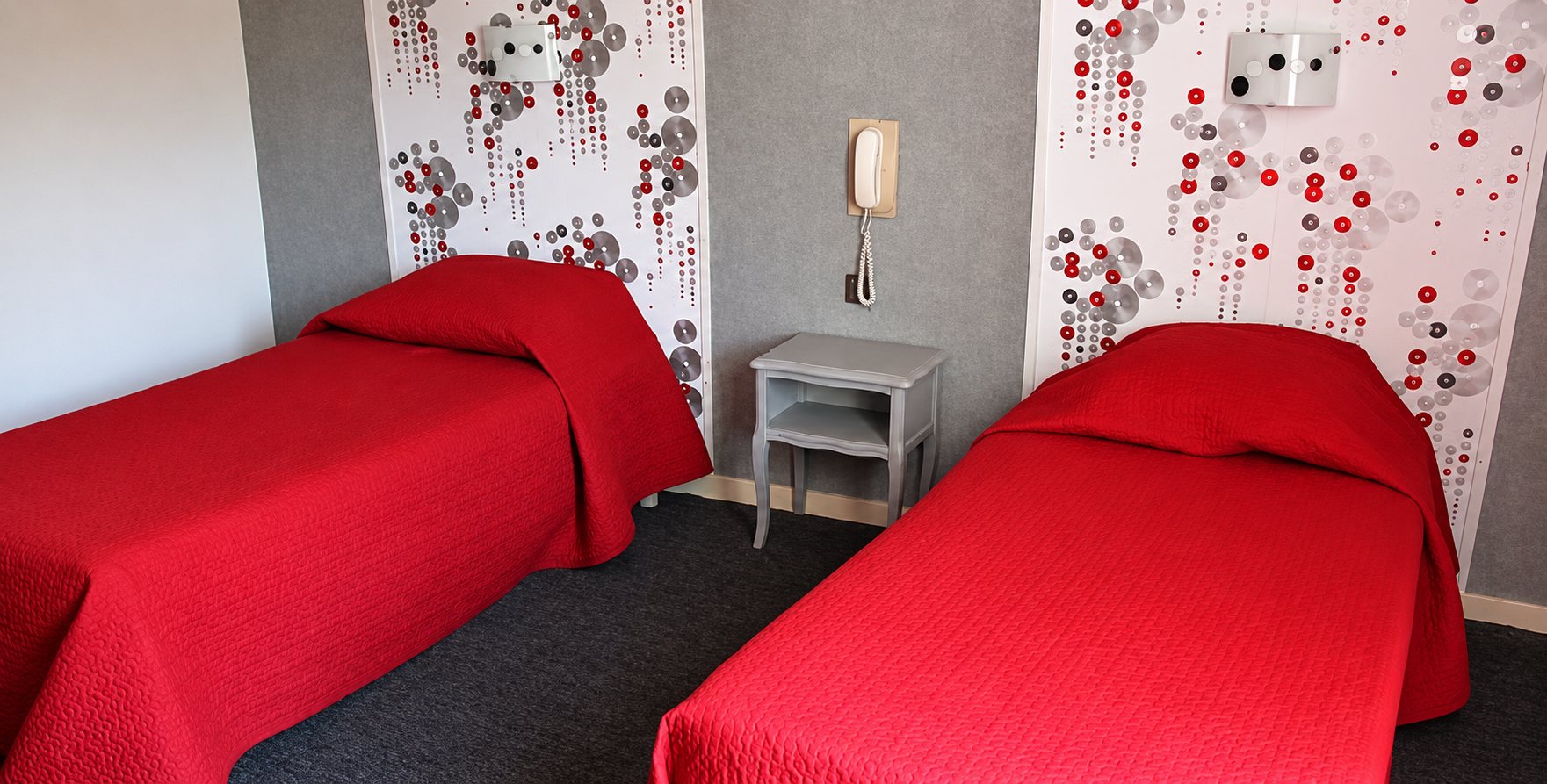 Chambre à deux lits simples recouverts de couvre-lits rouges, séparés par une table de chevet blanche avec téléphone mural, devant un papier peint à motifs de cercles rouges.