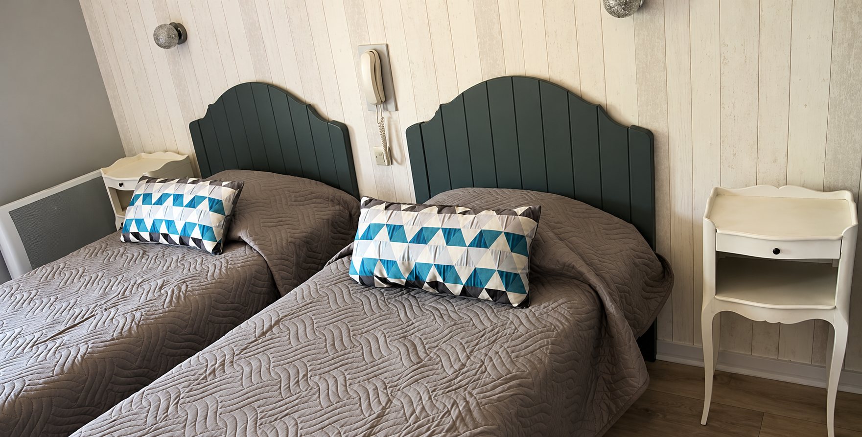 Chambre avec deux lits jumeaux couverts de couvre-lits gris matelassés, têtes de lit vert foncé, coussins à motifs bleus et tables de chevet blanches.