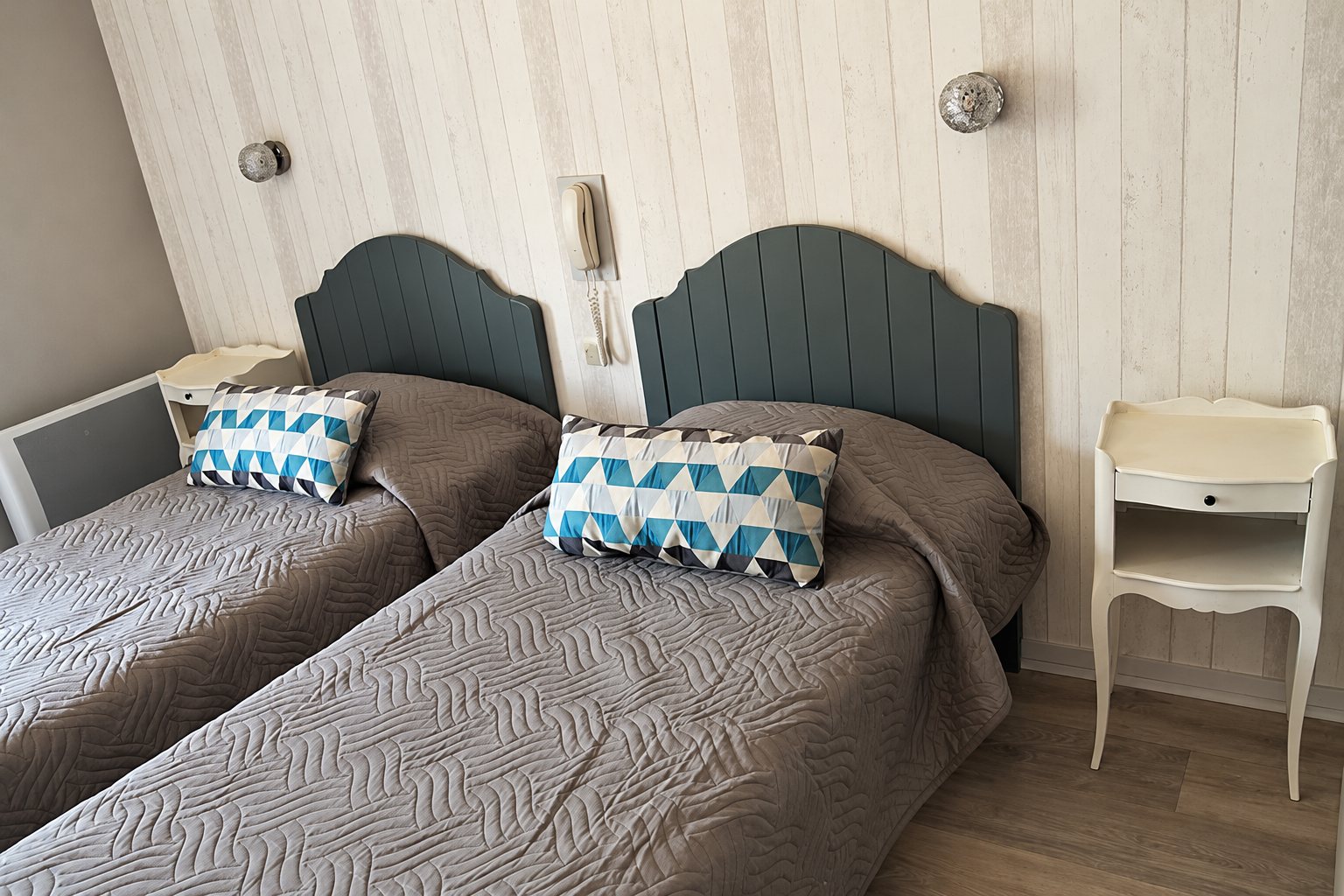 Chambre avec deux lits aux couvre-lits gris matelassés, têtes de lit vert-gris, coussins à motifs bleus et tables de chevet blanches.