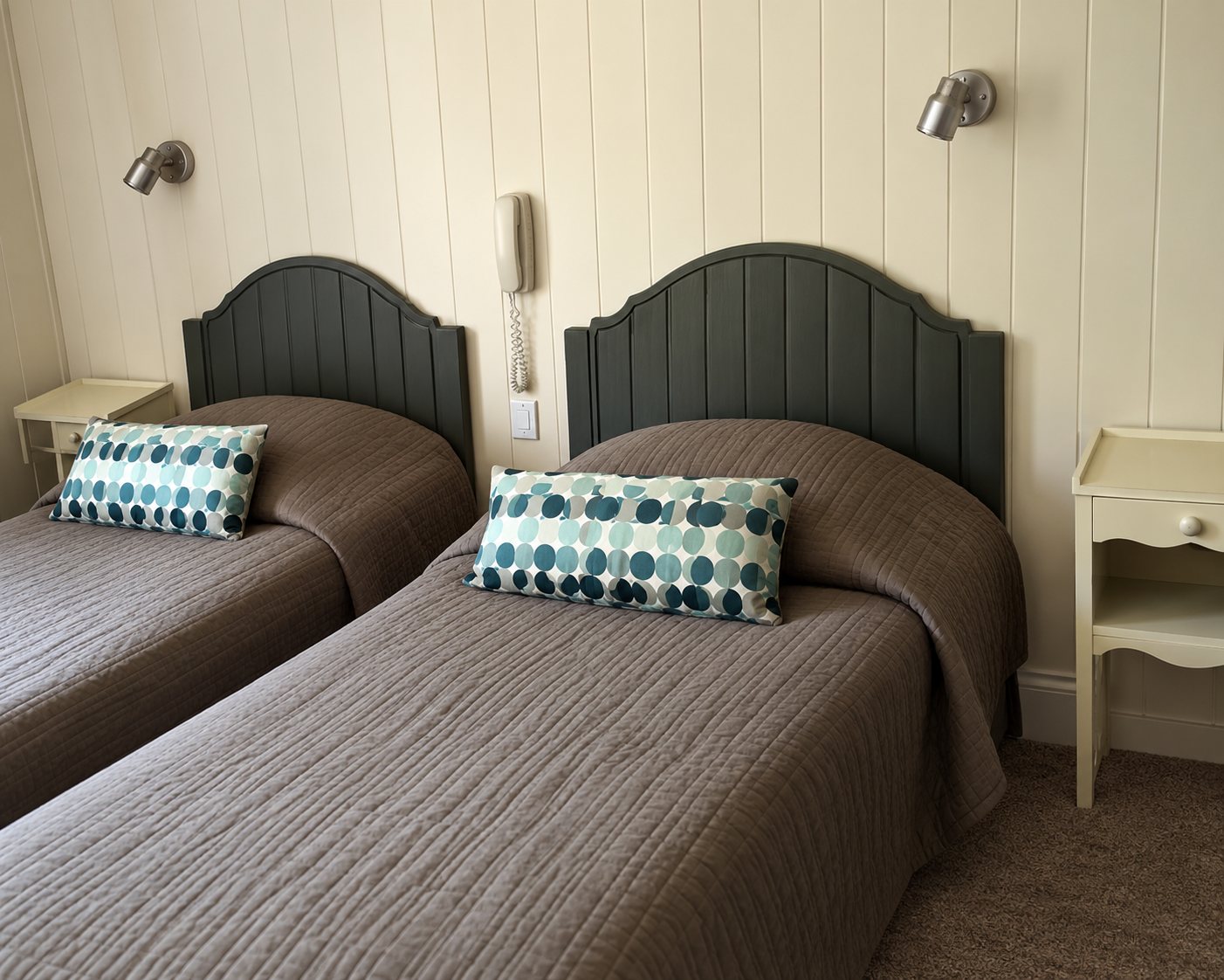 Une chambre d'hôtel meublée de deux lits jumeaux aux têtes de lit grises, ornés de coussins turquoise, avec des lampes murales au-dessus.