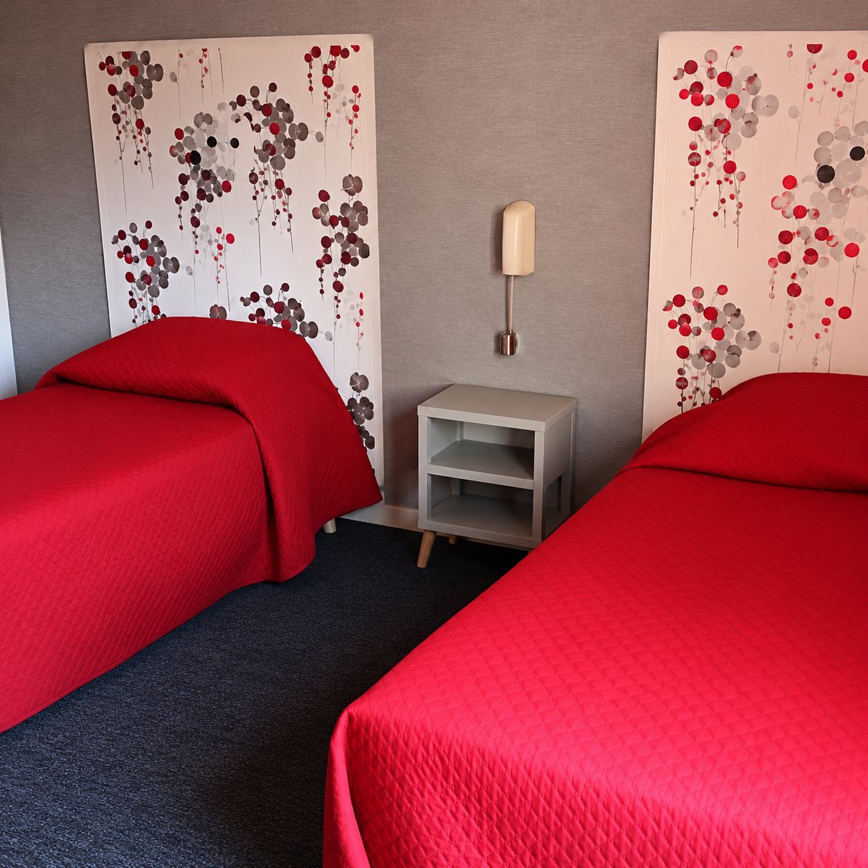 Chambre avec deux lits aux couvre-lits rouges, séparés par une table de chevet avec une lampe, sous des panneaux muraux décorés de motifs floraux.