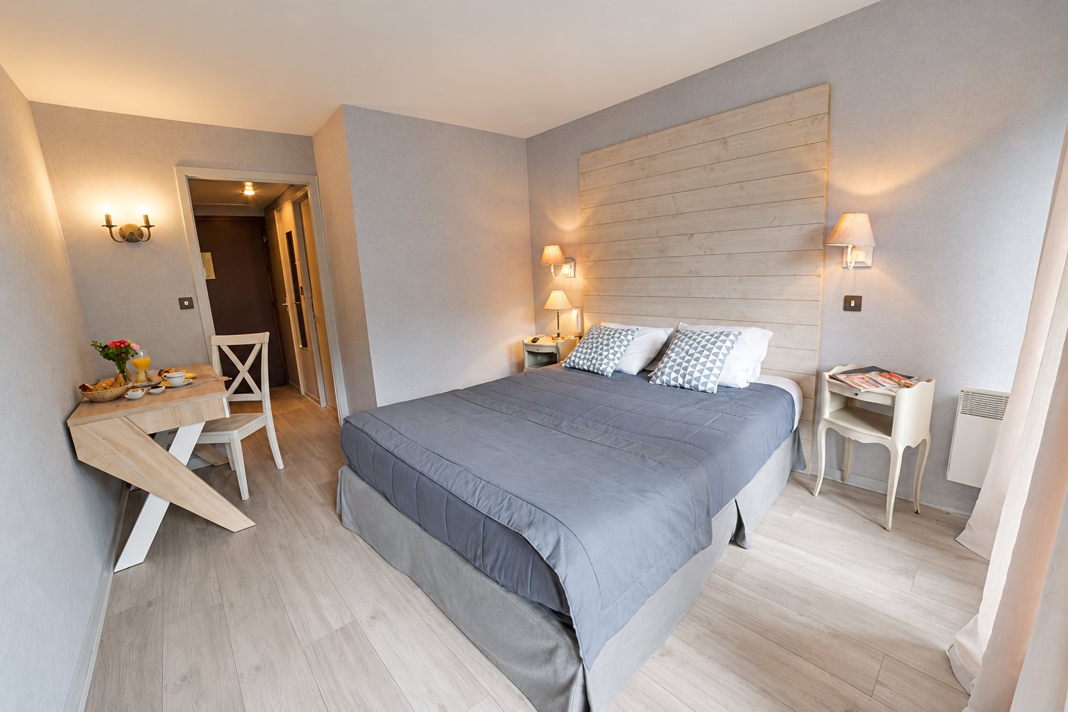 Chambre d'hôtel aux tons gris et blancs avec un grand lit double, une tête de lit en lattes de bois clair, des appliques murales et un petit bureau avec chaise.