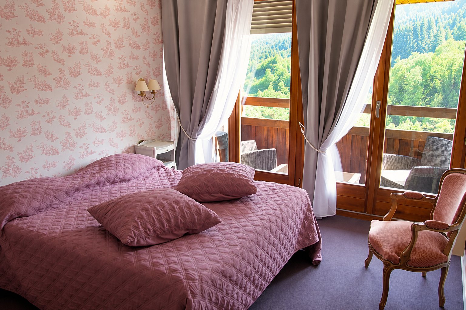 Chambre d'hôtel lumineuse avec un grand lit couvert d'un dessus-de-lit mauve matelassé, des rideaux gris, un fauteuil rose et une porte-fenêtre ouvrant sur un balcon en bois.