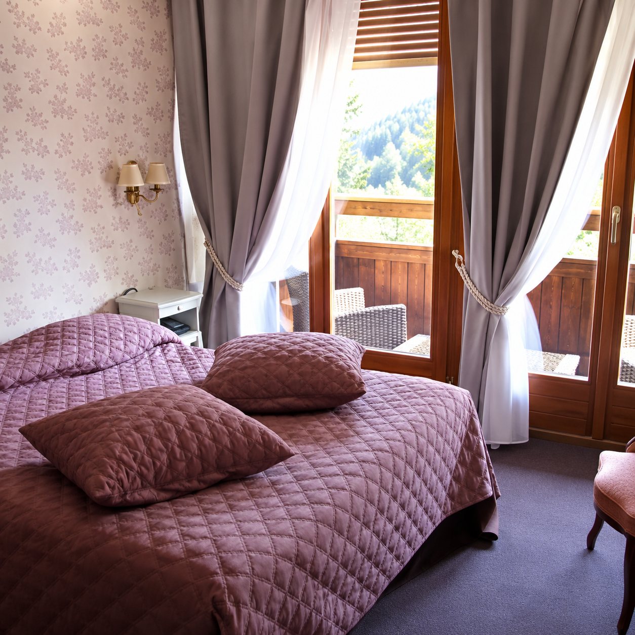 Une chambre d'hôtel lumineuse avec un lit couvert d'un dessus-de-lit rose matelassé, des rideaux gris et une porte-fenêtre donnant sur un balcon en bois.