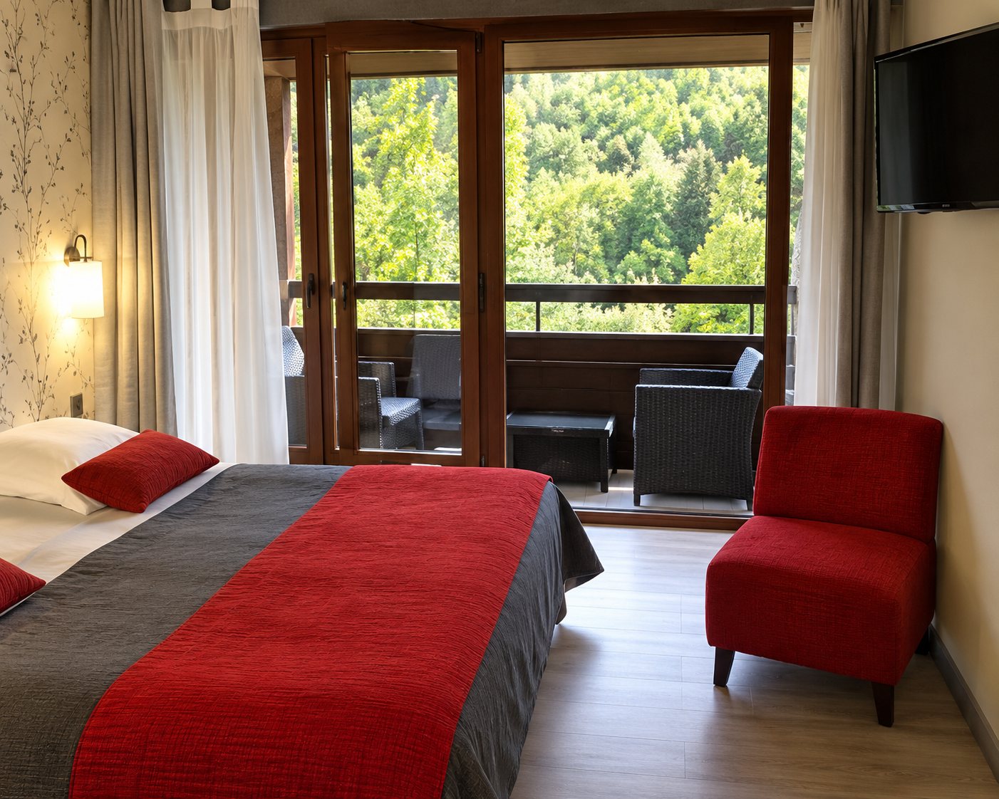 Chambre d'hôtel lumineuse avec un lit couvert d'un plaid rouge, un fauteuil rouge et une grande porte-fenêtre donnant sur la verdure.