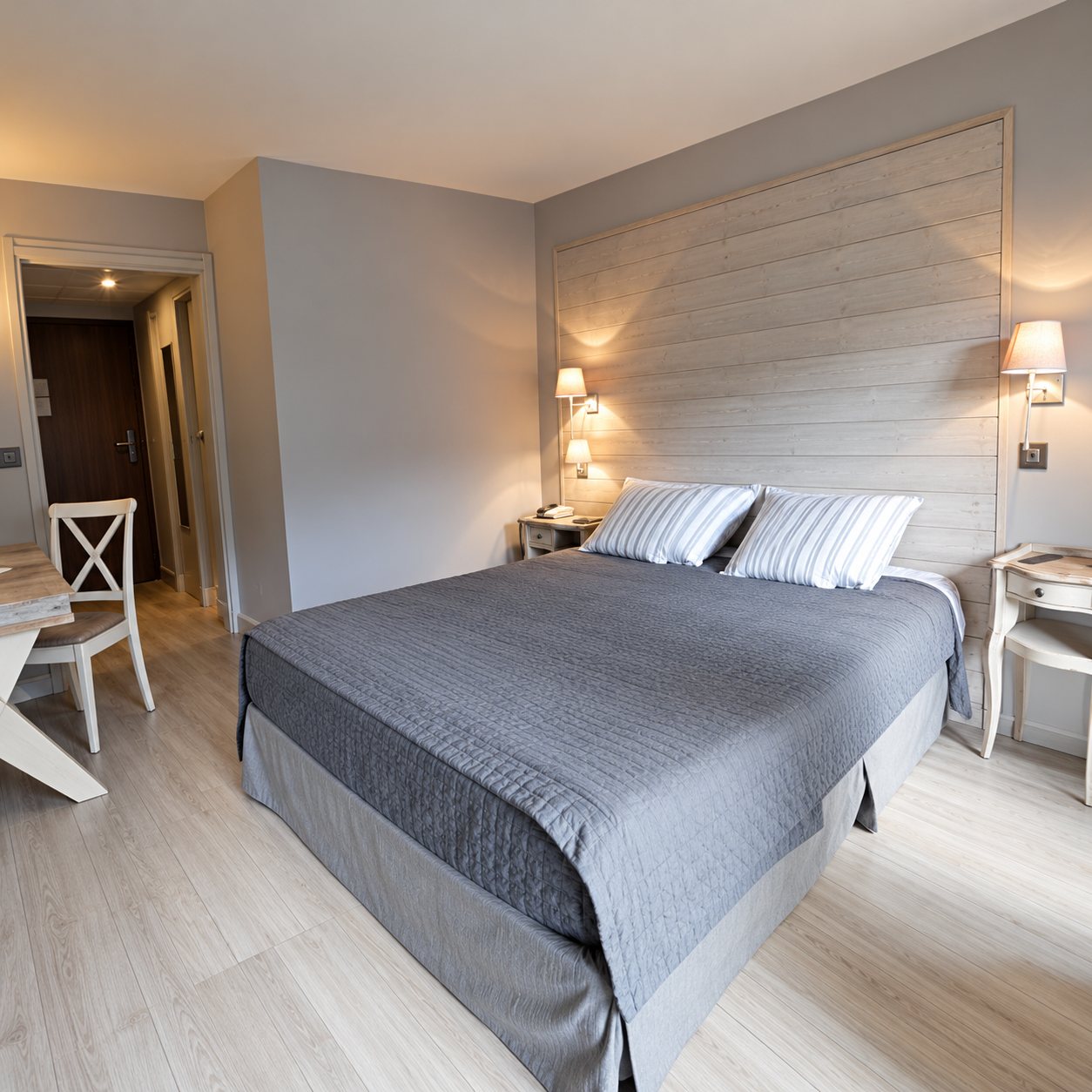 Chambre d'hôtel aux tons clairs avec un grand lit couvert d'un plaid gris, des lampes murales allumées et une chaise blanche près de l'entrée.