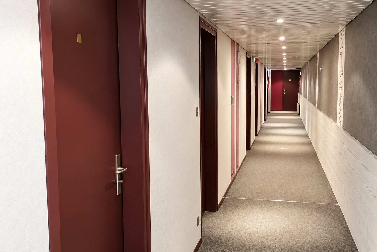 Un long couloir intérieur éclairé par des spots au plafond, bordé de portes bordeaux et de murs clairs, avec une moquette grise au sol.