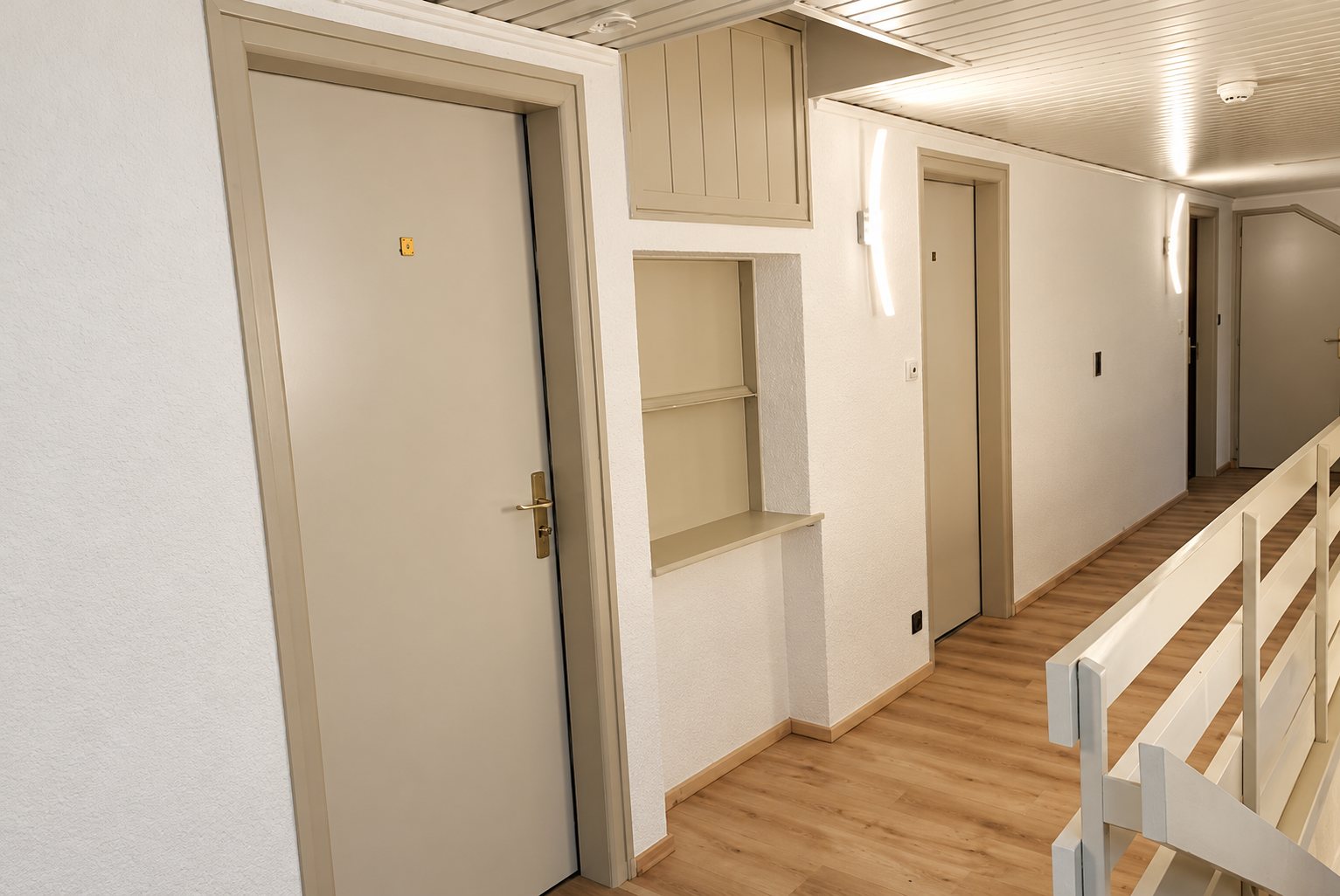 Couloir lumineux fraîchement rénové avec des portes d'appartement beige clair, un parquet clair, des appliques murales et une rampe d'escalier blanche.