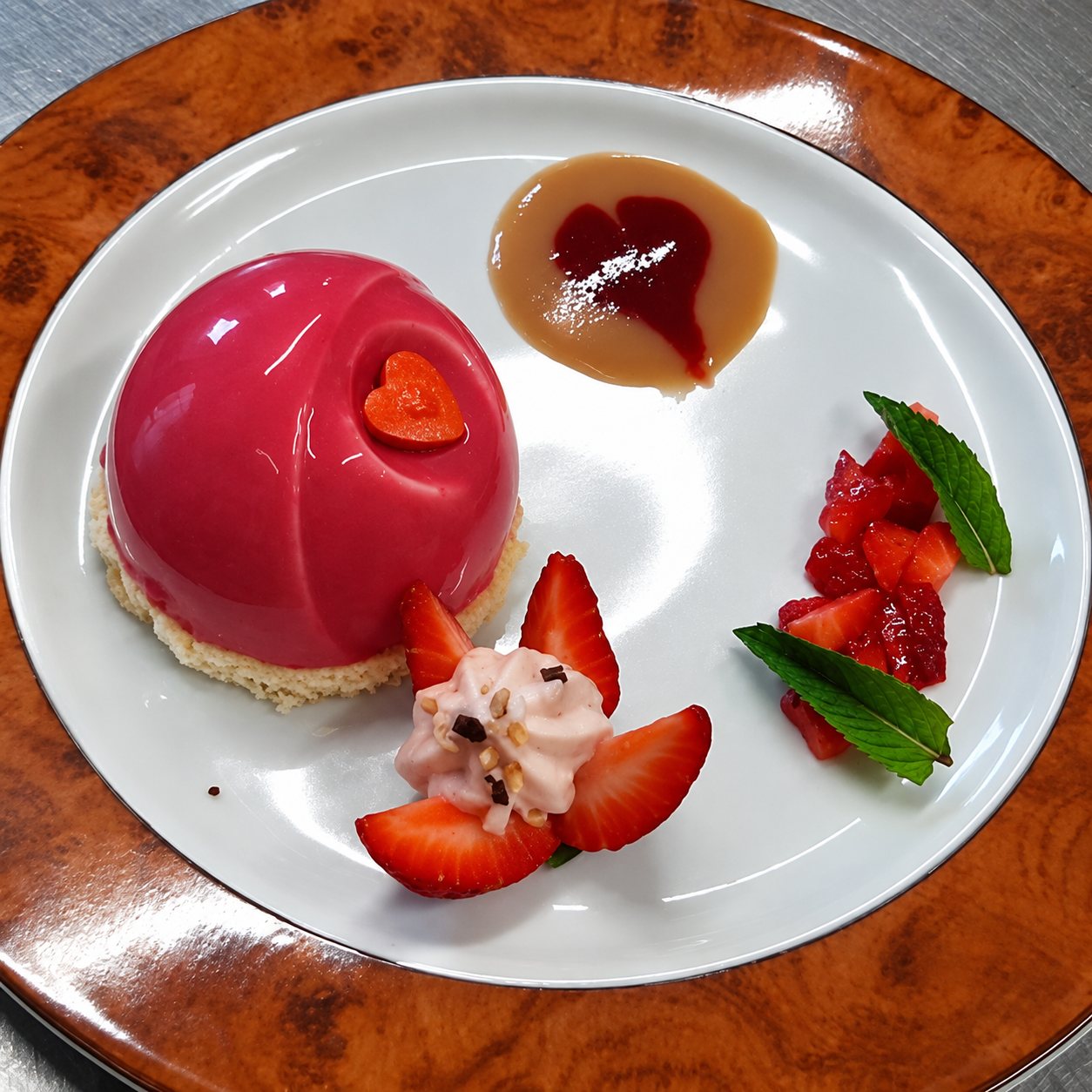 Assiette blanche garnie d'un dôme rose glacé orné d'un cœur, de fraises fraîches, d'une crème et de feuilles de menthe, posée sur un plateau en bois ronceux.