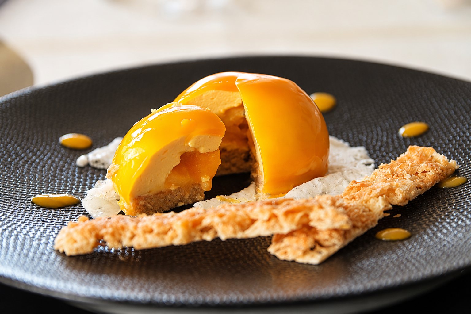 Un dessert en forme de dôme nappé de glaçage orange, coupé en deux, servi sur une assiette texturée noire avec des tuiles croustillantes et des gouttes de coulis.