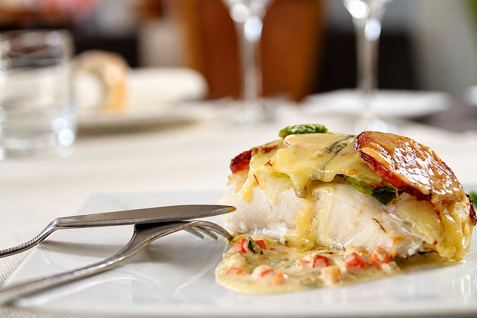 Un filet de poisson blanc nappé de sauce beurre blanc et garni de fines tranches de chorizo, présenté sur une assiette blanche avec des couverts, dans un restaurant.