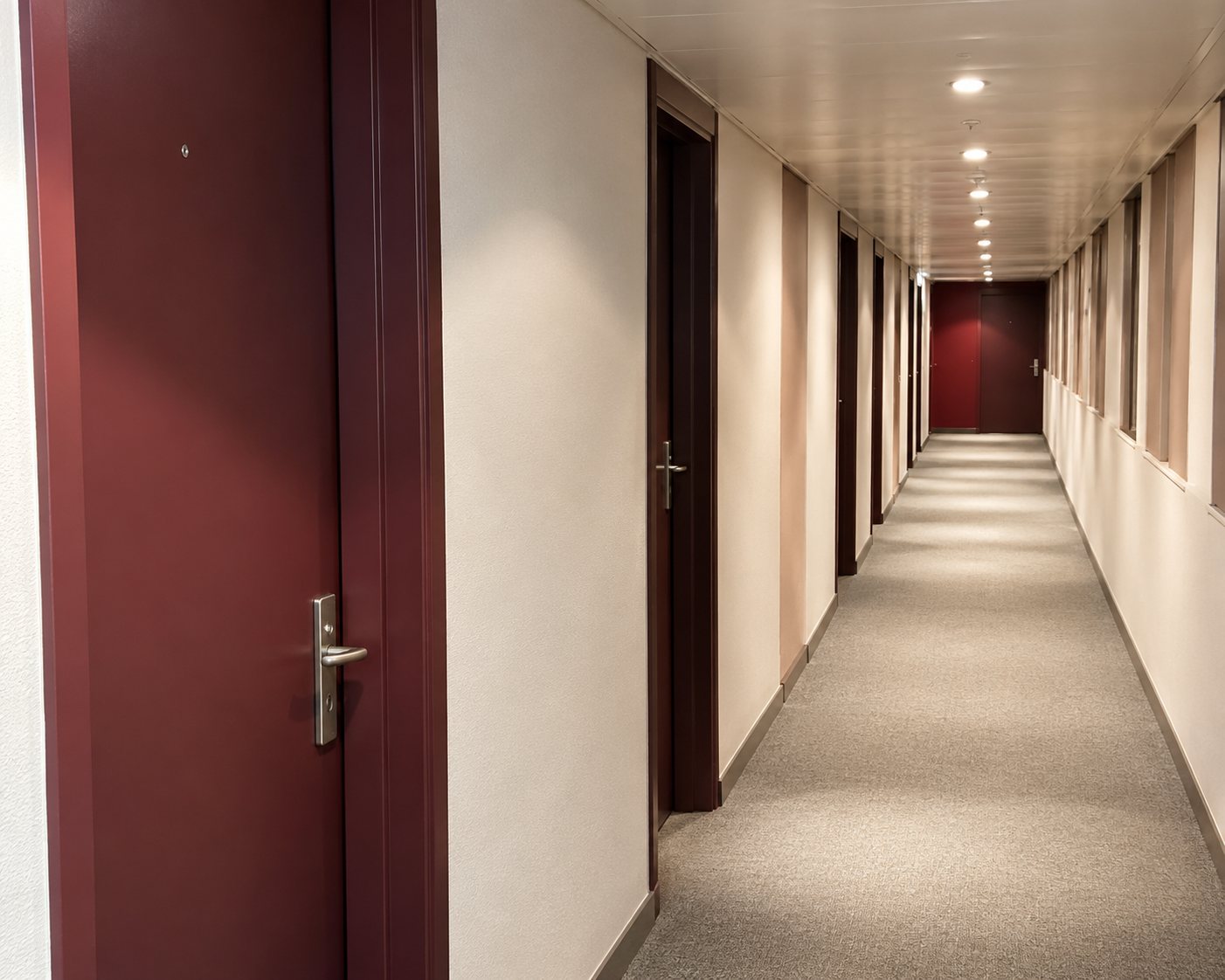 Un long couloir étroit aux murs clairs et aux portes en bois sombre, éclairé par des spots au plafond et menant à une porte rouge au fond.