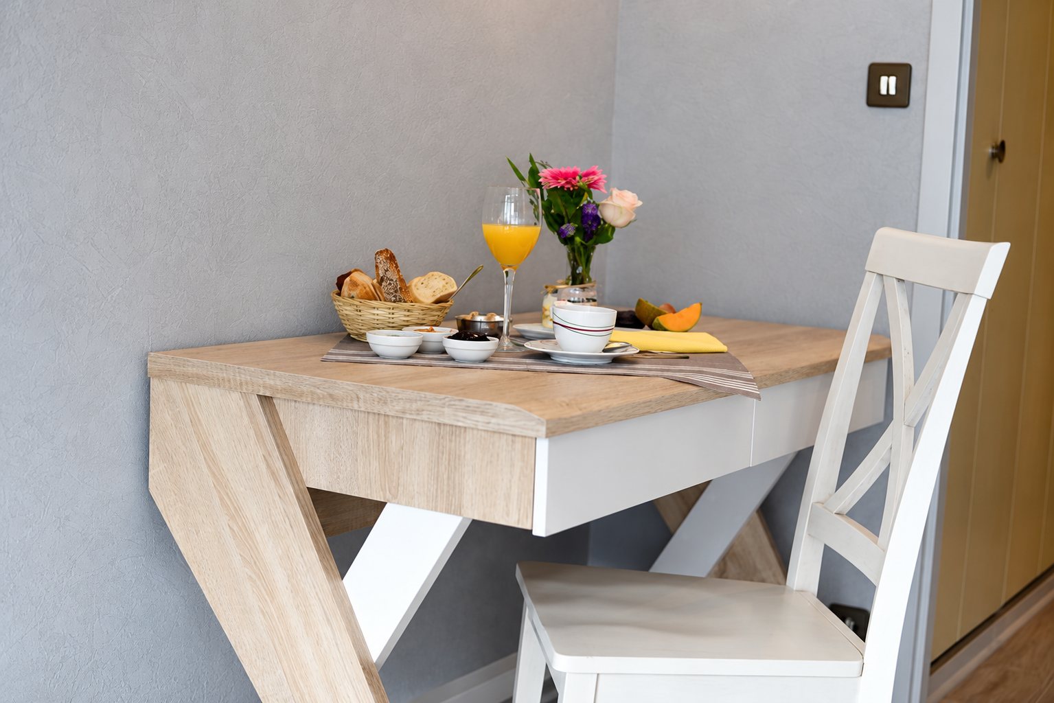 Un plateau de petit-déjeuner avec viennoiseries, jus d'orange, fruits et fleurs est posé sur une petite table en bois clair, à côté d'une chaise blanche.