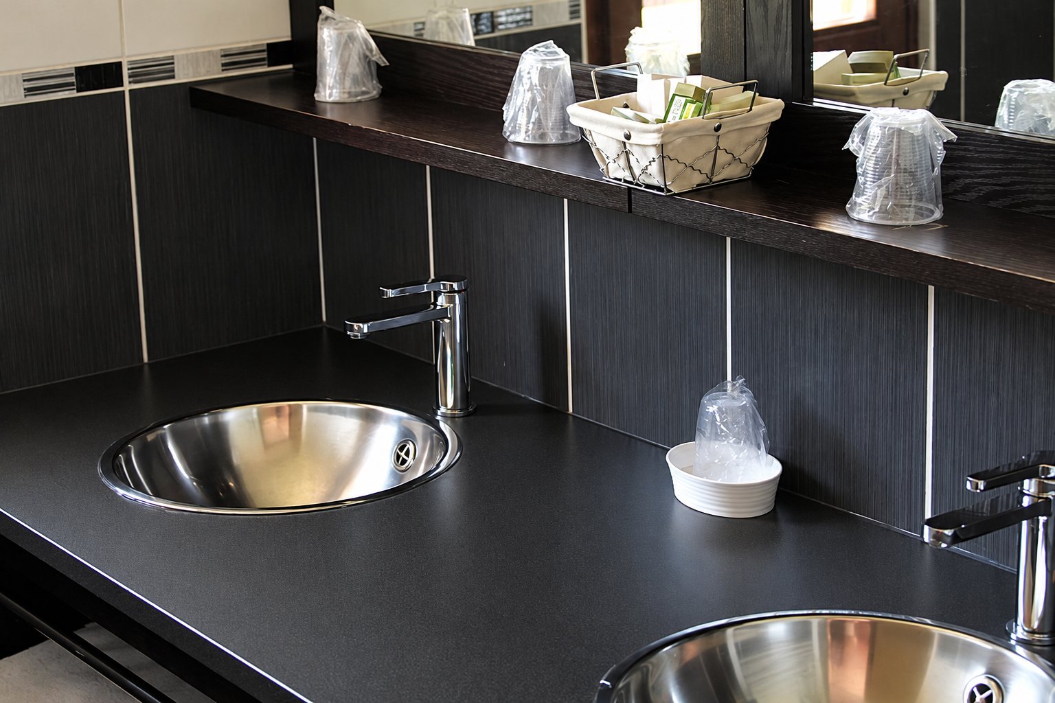 Un plan de toilettes noir équipé de deux vasques rondes en inox, de robinets chromés et de gobelets en plastique posés sur une étagère devant un miroir.