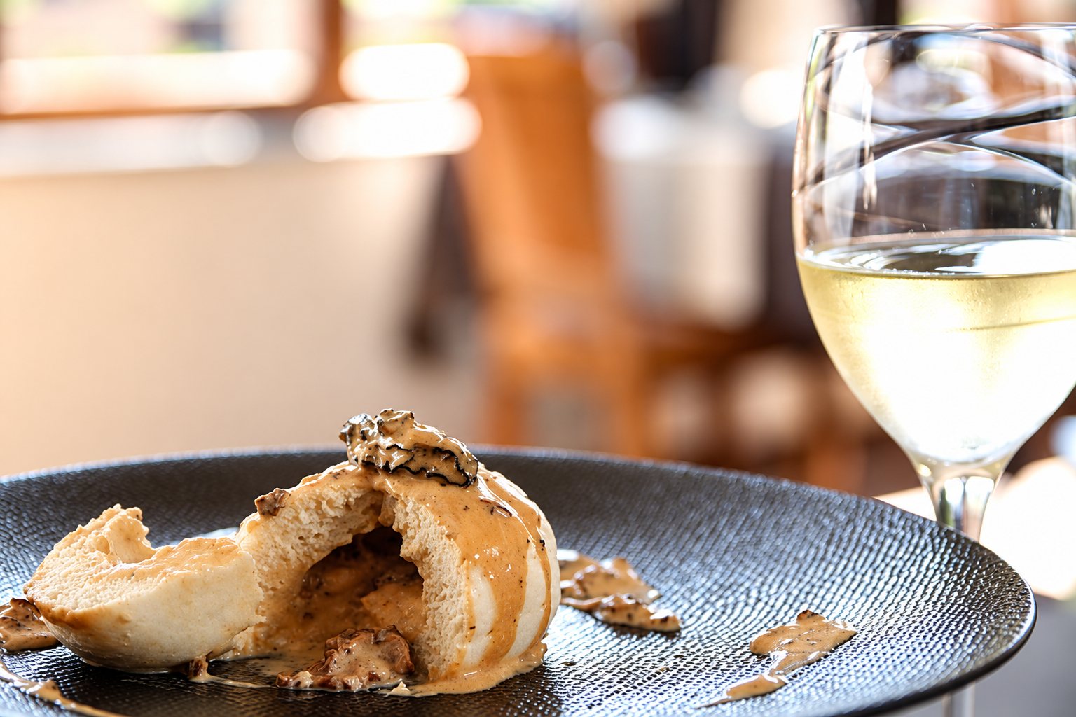 Un plat nappé de sauce crémeuse sur une assiette noire, avec un verre de vin blanc.