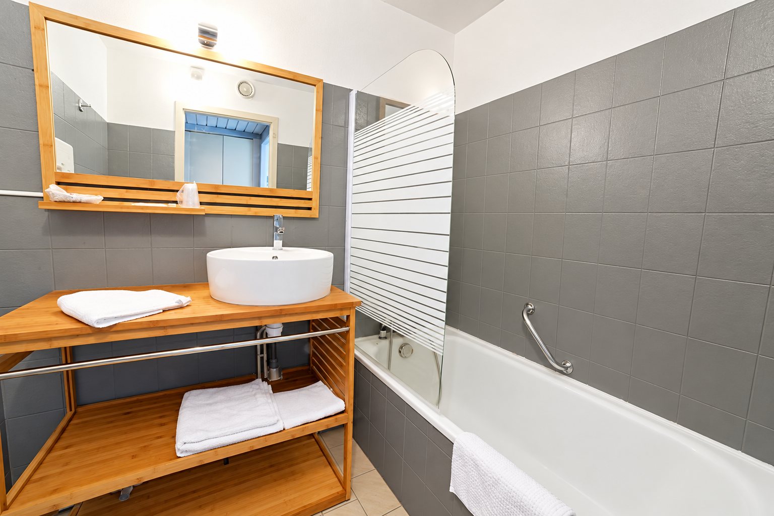 Salle de bain aux carreaux gris avec meuble en bambou, vasque blanche ronde, grand miroir et baignoire avec pare-douche à rayures.