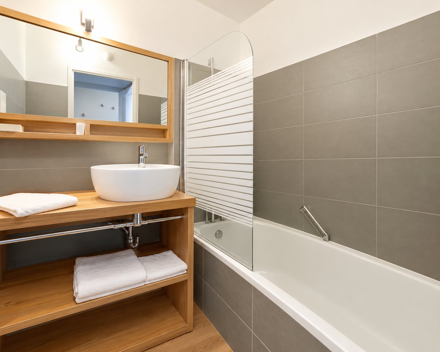 Salle de bains lumineuse avec un meuble vasque en bois clair, un grand miroir et une baignoire avec pare-baignoire en verre.