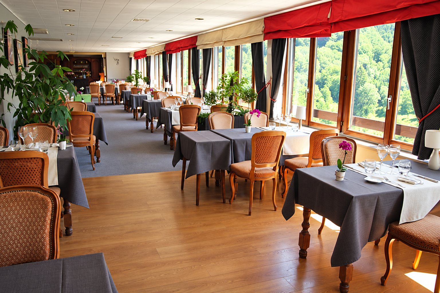 Une salle de restaurant aux tables dressées avec nappes grises et chaises capitonnées, éclairée par de larges fenêtres donnant sur une vallée boisée.
