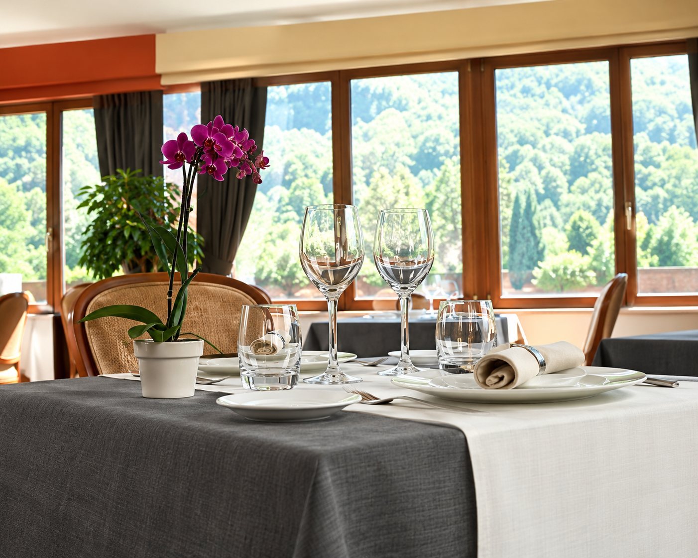 Intérieur d'un restaurant avec des tables dressées de verres et de nappes sombres, face à de grandes baies vitrées donnant sur la verdure.