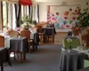 Intérieur d'une salle de restaurant avec plusieurs tables dressées de nappes grises et des chaises en bois devant de grandes fenêtres.