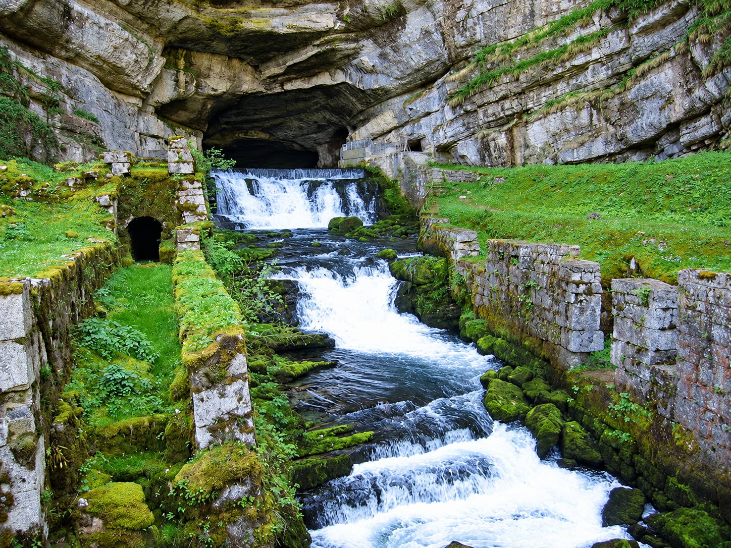 La source de la Loue sortant d'une grotte et dévalant en cascades entre des murs de pierre couverts de mousse.