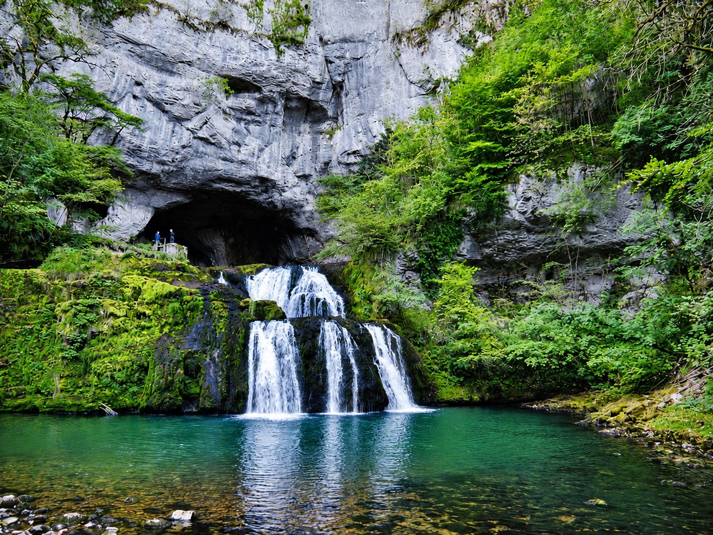 La source du Lison : des cascades tombent d'une grotte dans un bassin vert, au pied d'une paroi rocheuse.