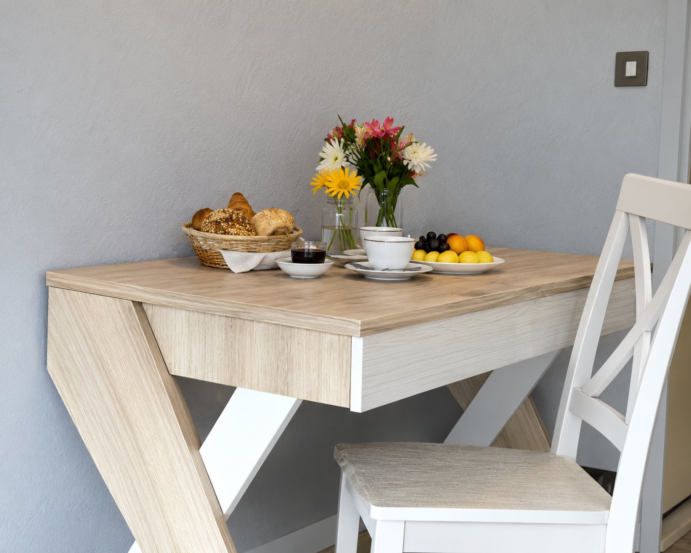 Une table en bois clair aux pieds en forme de Z, dressée avec des fleurs dans un vase, accompagnée d'une chaise blanche devant un mur clair.