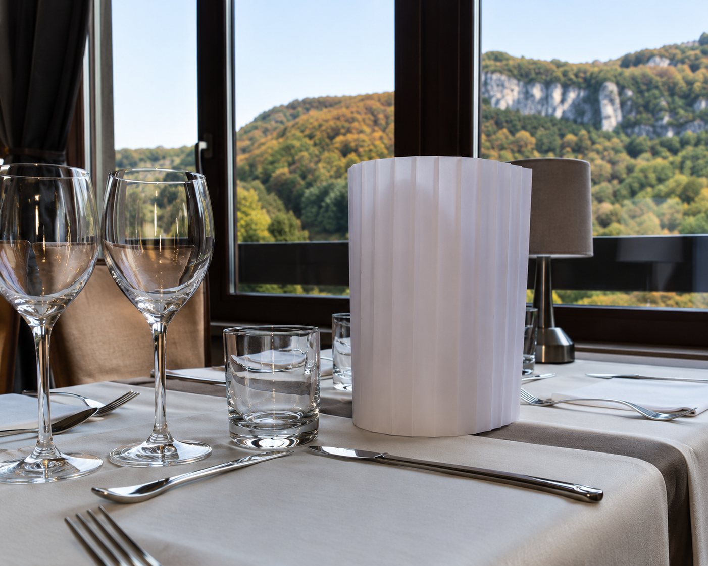 Une table de restaurant dressée avec des verres à pied et un photophore blanc, devant une grande fenêtre donnant sur l'extérieur.