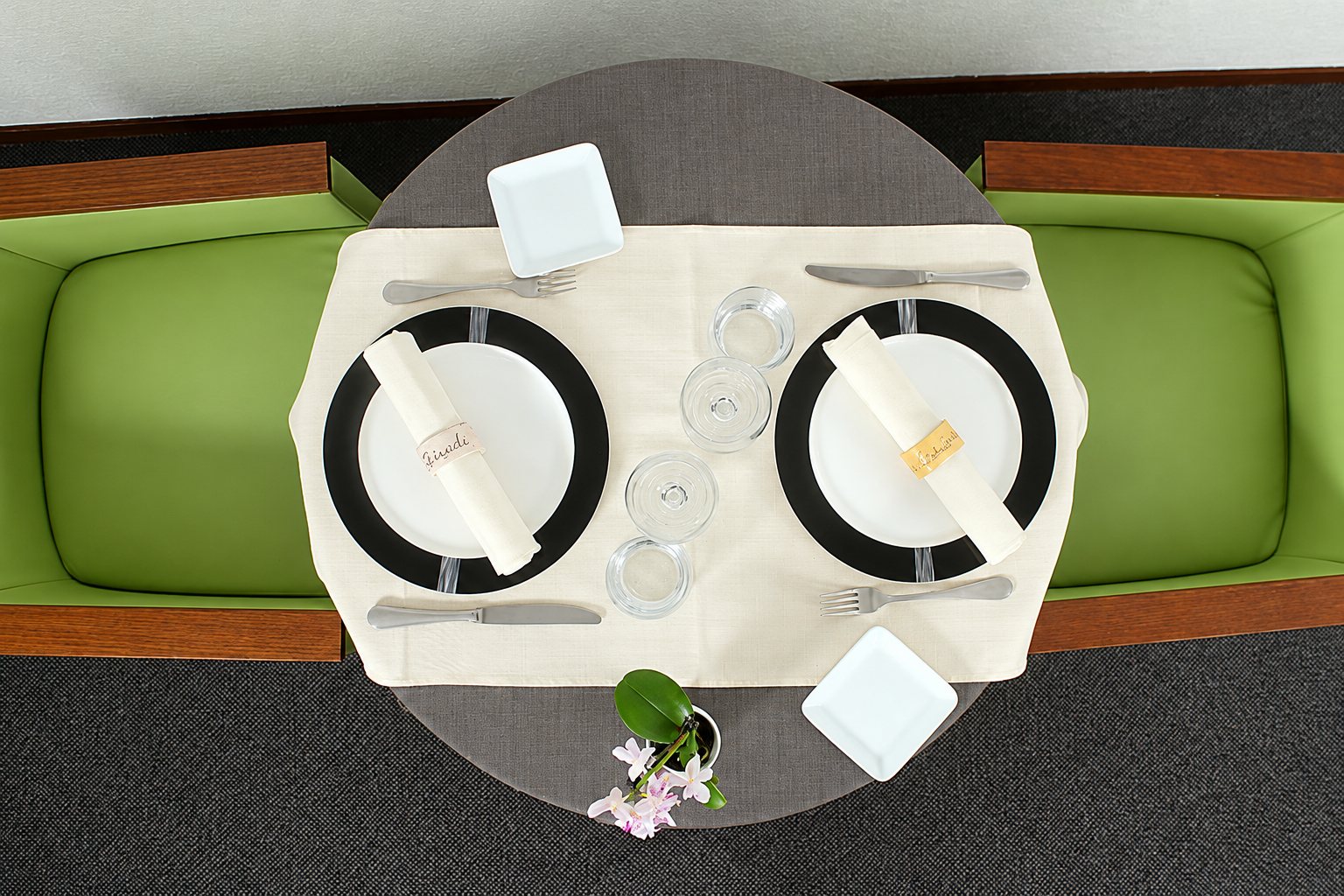 Vue de dessus d'une table ronde dressée pour deux personnes avec assiettes noires, serviettes blanches, verres et une petite orchidée, entre deux banquettes vertes.