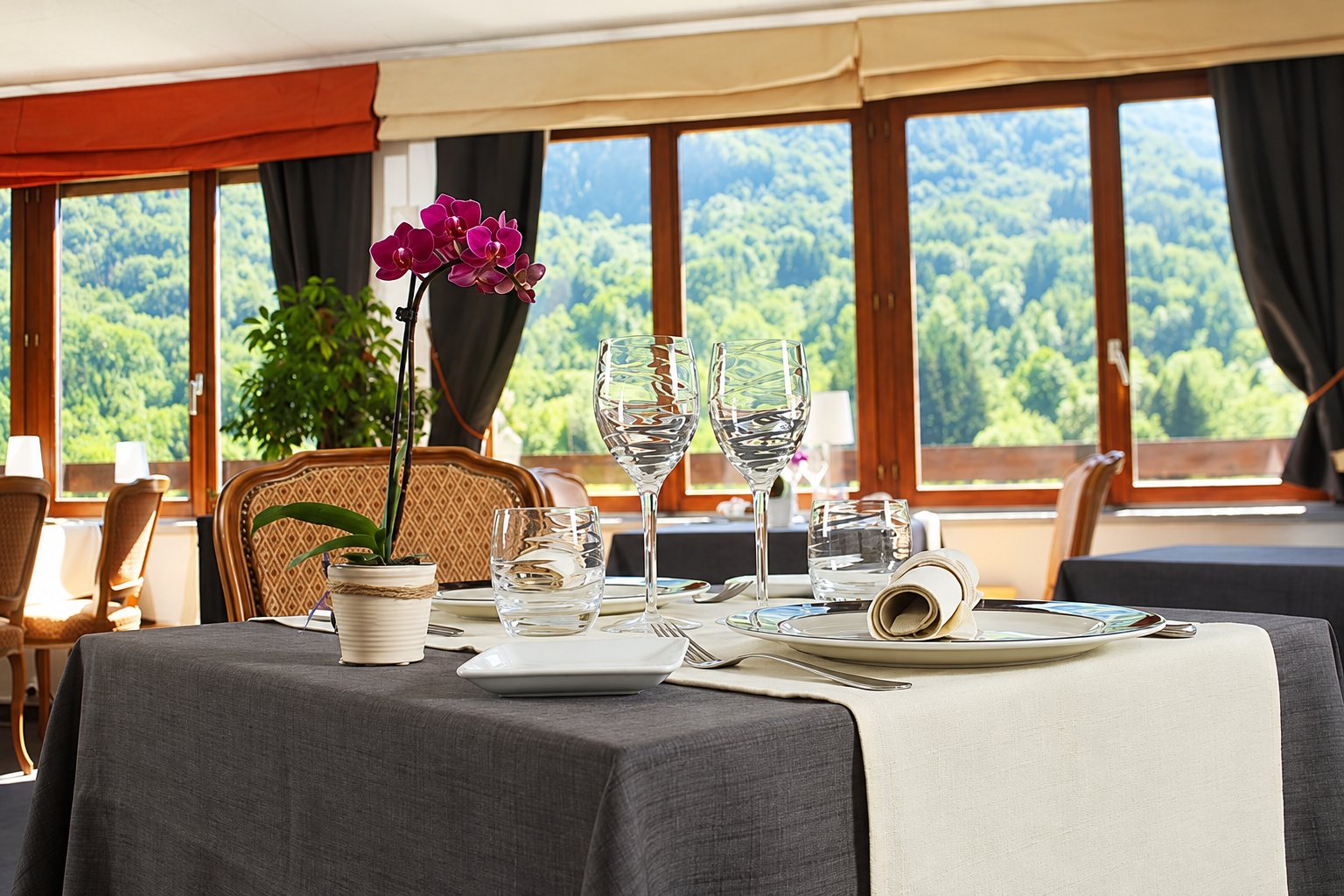 Table de restaurant dressée avec nappe grise, verres et orchidée rose, devant de grandes fenêtres donnant sur une colline boisée.